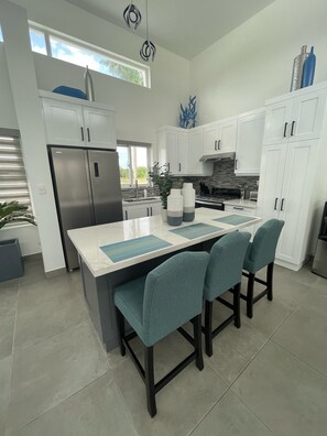 A modern kitchen with white cabinets, a stainless steel refrigerator, a central island with chairs, and a view of the outdoors. - Sosua Ocean Village Luxury Villa: 3BR/3.5BA Pool Oasis (Sosúa)