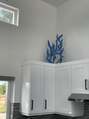 Cuina amb armaris blancs, un objecte decoratiu blau a la paret i vista a vegetació a l'exterior.
