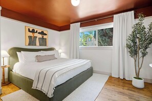 Ein Schlafzimmer mit einem Bett, einem Fenster mit Vorhängen, einer Topfpflanze und einem Bild an der Wand.