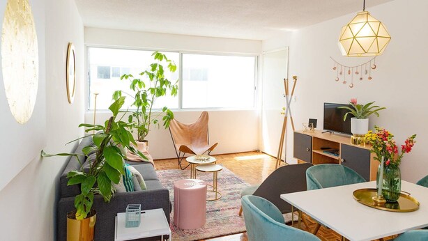 Ein modernes Wohnzimmer mit einer Couch, Sesseln, einem Couchtisch und einem Esstisch. Ein Fernseher steht auf einem Schrank, eine Pflanze und eine Hängeleuchte sind ebenfalls vorhanden.
