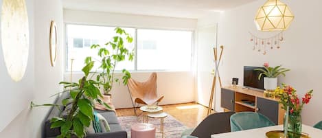 A modern living room with a sofa, chairs, a coffee table, and a dining table. There is a television on a stand, a plant, and a hanging light fixture.
