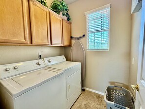 Un espace buanderie comprenant un lave-linge et un sèche-linge, des armoires en bois, une fenêtre avec des stores et un panier à linge.