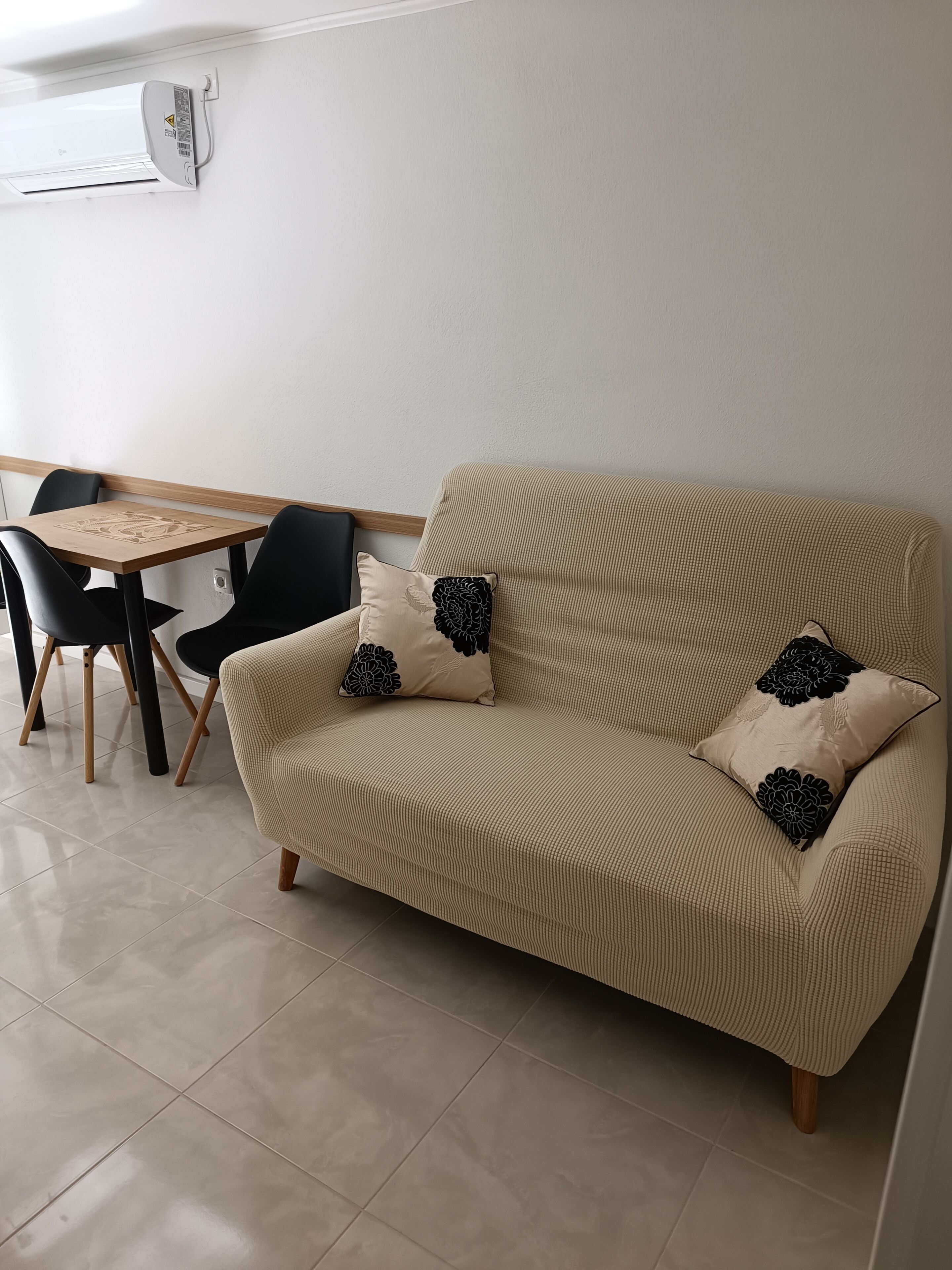 A beige sofa with black floral cushions, a wooden table with chairs, and a white wall with an air conditioning unit.