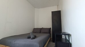 A single bed with a grey mattress and two pillows, a black nightstand with a small table, and a tall black cabinet against a plain wall.