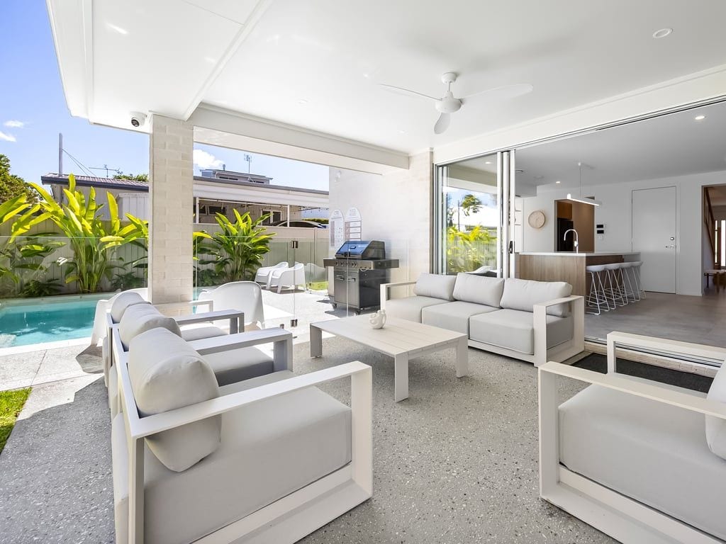 Uma área de estar moderna ao ar livre com piscina, móveis brancos e uma porta de correr de vidro que dá para uma cozinha.
