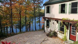 A house with a gravel patio, a red door, and a view of a lake surrounded by trees. - Stunning Lakefront Lodge with Dock and Hot Tub (Raymond)