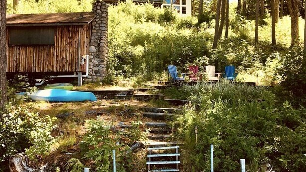 A rustic cabin with a stone chimney, surrounded by dense forest. - Stunning Lakefront Lodge with Dock and Hot Tub (Raymond)