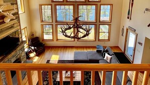 A cabin interior with a wooden ceiling, a fireplace, a large window with a deer head decoration, and a view of the outdoors. - Stunning Lakefront Lodge with Dock and Hot Tub (Raymond)