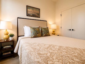 A neatly made bed with patterned bedding, two bedside lamps, a nightstand with books, and a painting on the wall. - The Jardin Bloom Suite | Near Downtown (Savannah)
