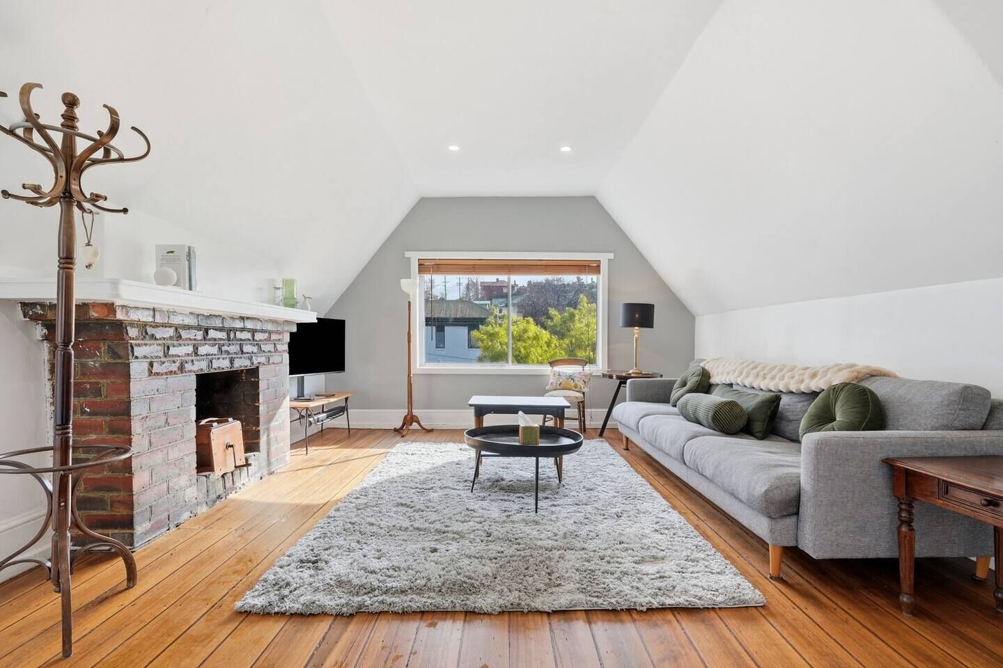 Ein Loft-Wohnzimmer mit Kamin aus Ziegelsteinen, einem grauen Sofa und einem großen Fenster mit Blick auf Grünflächen.