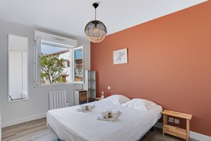 A bedroom with a bed, a bedside table, a nightstand, a radiator, a window, and a hanging light fixture. - Appartement Teilleria - Welkeys (Hendaye)