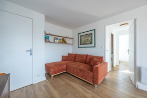 A modern living room with a sectional sofa, wooden flooring, a white door, a shelf with books, and a framed artwork. - Le Balcon du Grand Cap - vue mer - Welkeys (Villers-sur-Mer)