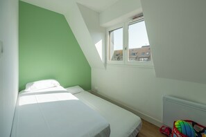 Ein kleines Zimmer mit einem Einzelbett, geneigter Decke und Fenster mit Blick auf ein Gebäude.