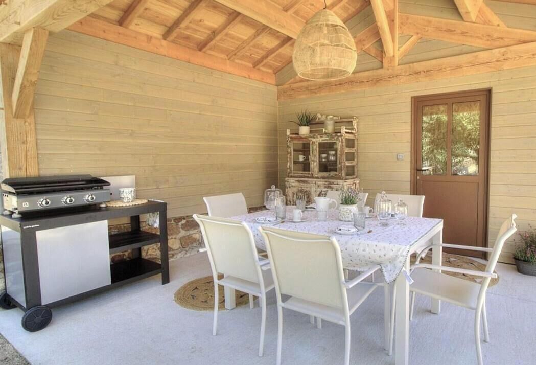 Uma área de refeições ao ar livre com churrasqueira, cadeiras brancas, mesa posta com copos e uma parede de madeira rústica.