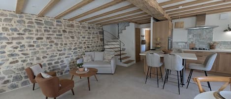 A spacious living area with a stone wall, wooden beams, a sofa, chairs, a dining table, and a kitchen in the background.