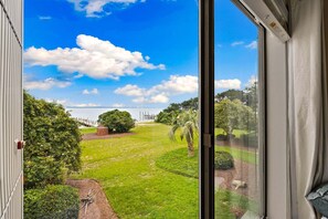 A view from a window overlooking a well-maintained lawn with a wooden deck and a body of water in the distance. - RSC203 - Wander Mint Soundfront condo with sunset views (Manteo)