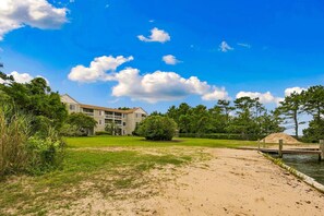 A building with multiple floors and balconies, a grassy area in front, and a body of water with a dock. - RSC203 - Wander Mint Soundfront condo with sunset views (Manteo)