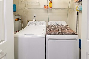 A laundry room with two washing machines, a water heater, and a shelf with cleaning supplies. - RSC203 - Wander Mint Soundfront condo with sunset views (Manteo)