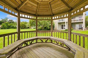 A wooden gazebo with a curved roof and open sides, surrounded by a grassy lawn and a white building with balconies. - RSC203 - Wander Mint Soundfront condo with sunset views (Manteo)
