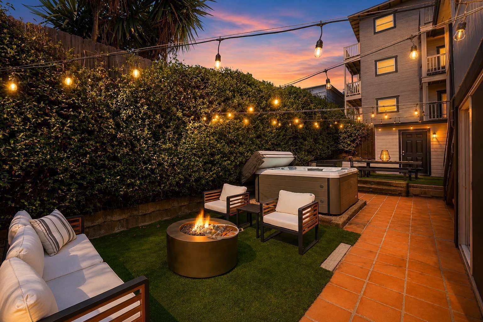 Une terrasse extérieure dotée d’un foyer, de sièges et d’un jacuzzi, le tout entouré d’une haie et illuminé de guirlandes lumineuses.