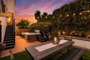 A patio with a hot tub, outdoor seating, and string lights against a backdrop of a hedge and a sunset sky. - Golden Hill 7BR Retreat with Private Hot Tub (San Diego)