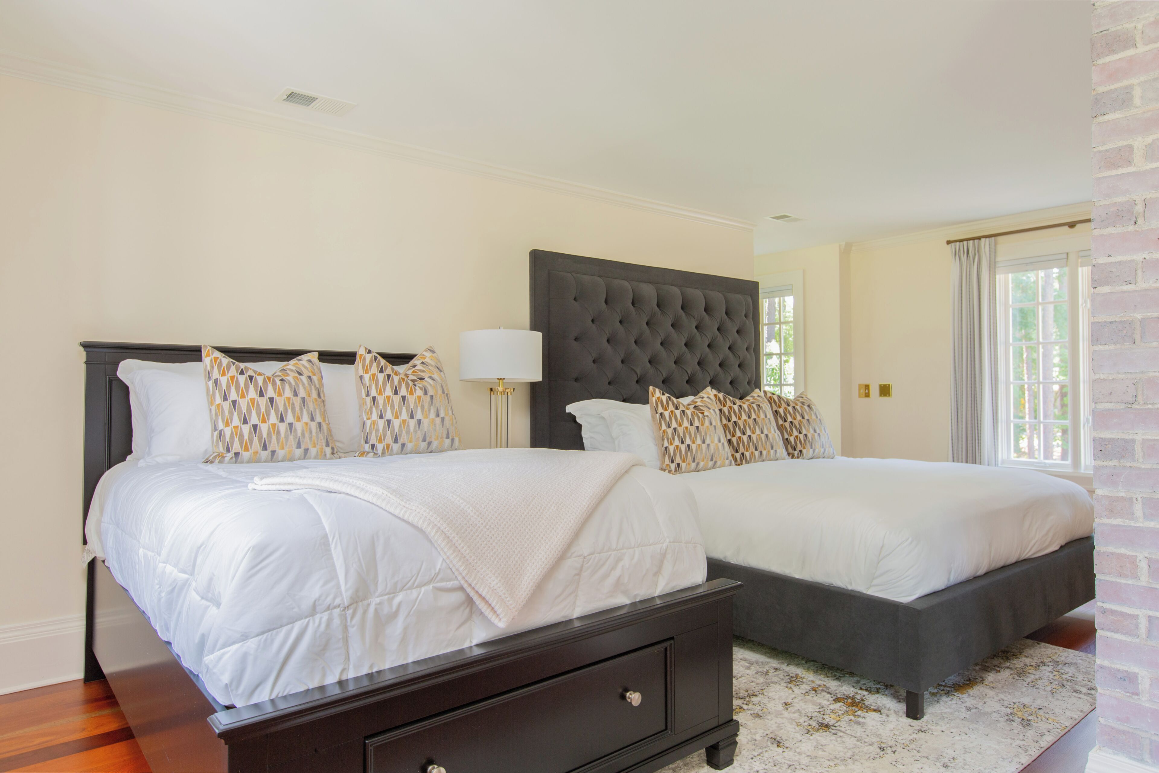 A double bed with a dark headboard and multiple pillows, a bedside table with a lamp, and a view of a brick wall and window.