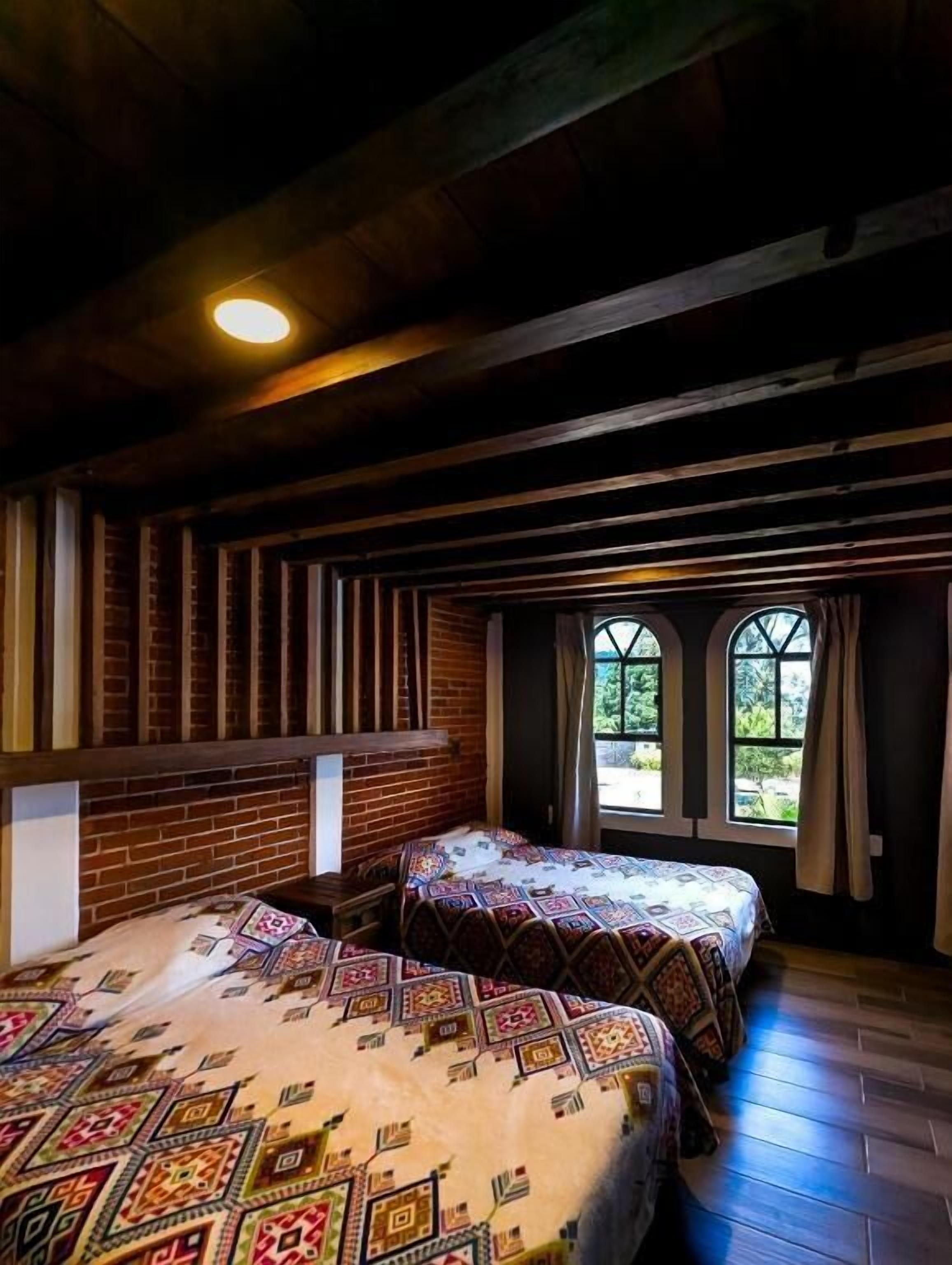 A room with two beds, wooden beams, brick walls, and windows with curtains.