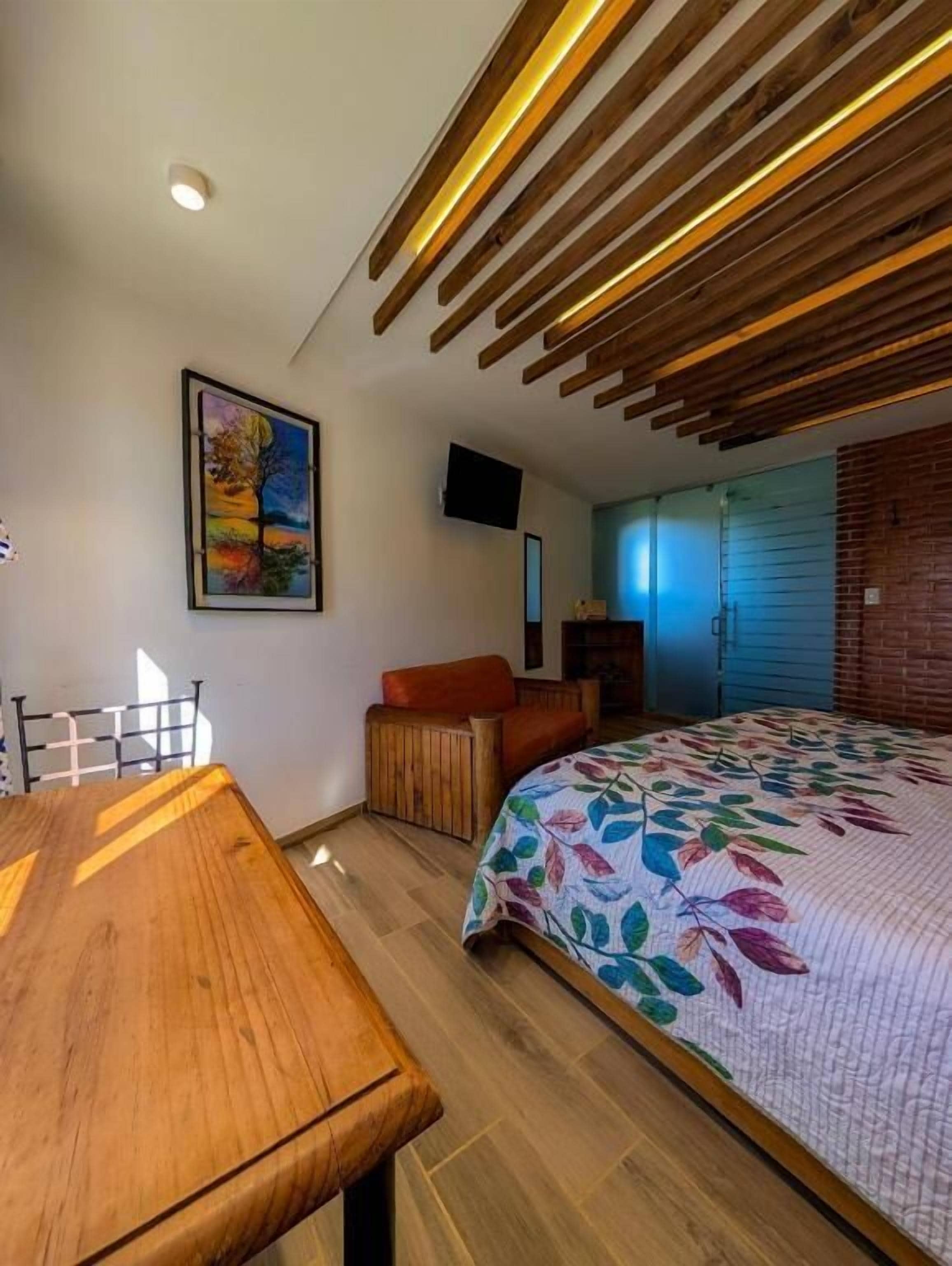 A bedroom with a wooden ceiling, a bed with a floral bedspread, a desk, and a chair.