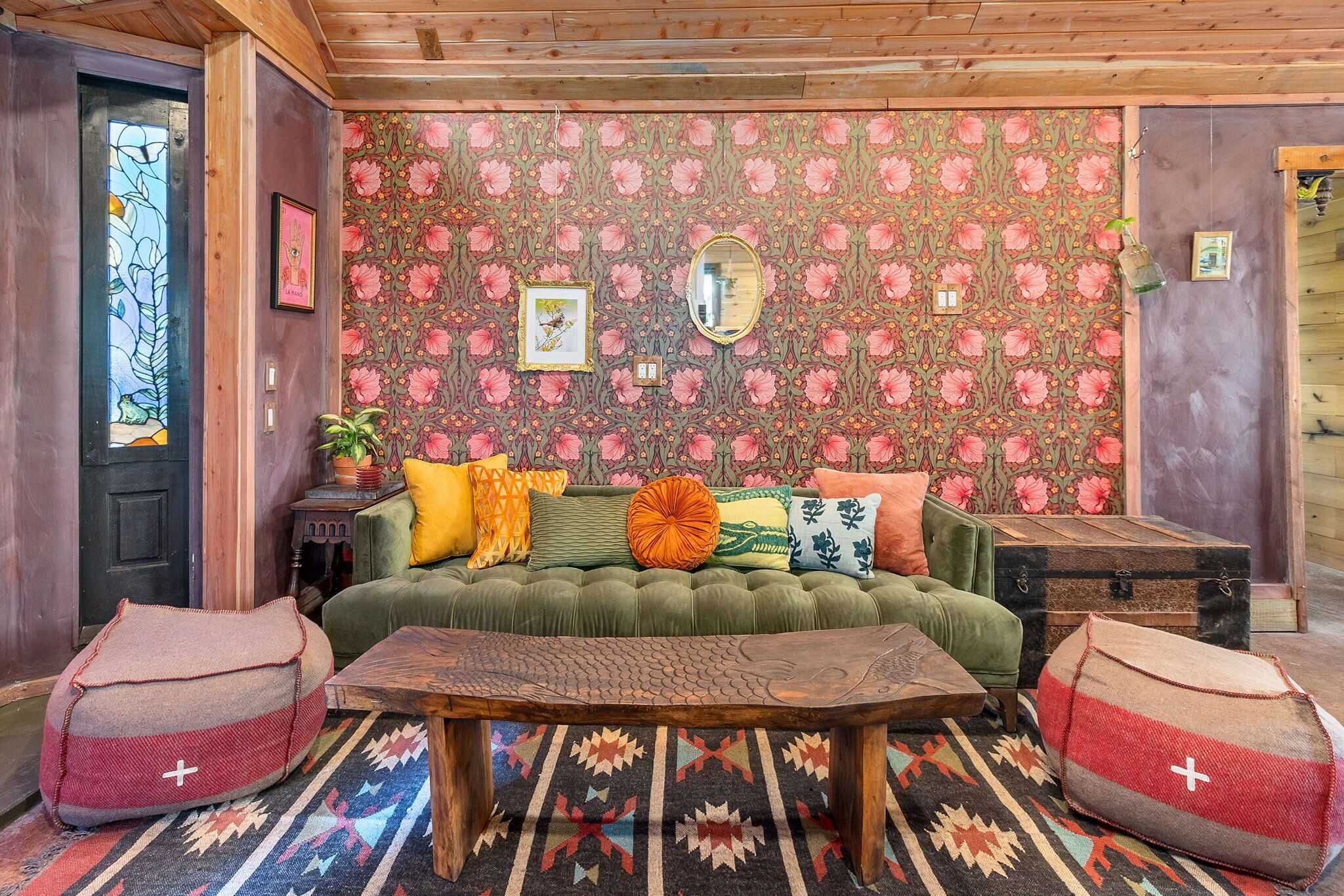 Un salon chaleureux avec un papier peint à motifs, un canapé vert, une table basse en bois et deux fauteuils pouf.