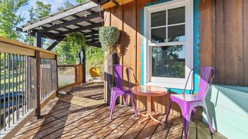 A wooden deck with a pergola, a window with a blue trim, and two purple chairs with a small table.