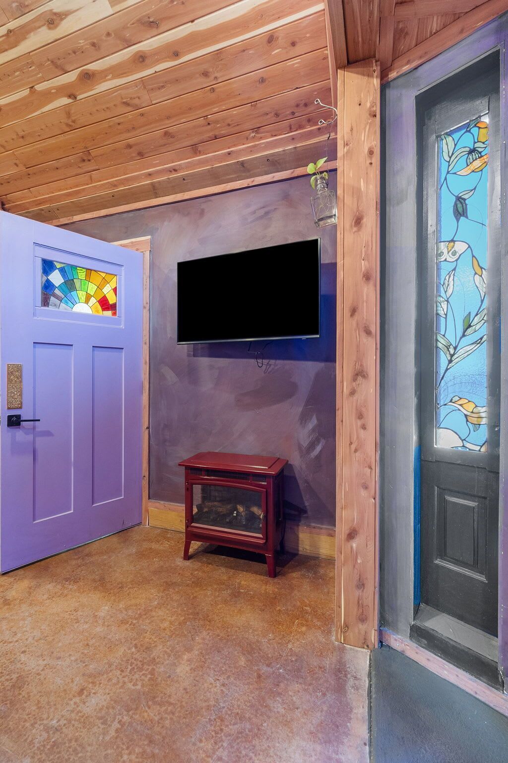 A room with a wooden ceiling, a purple door with a stained glass window, a black flat-screen TV mounted on the wall, and a small wooden table.