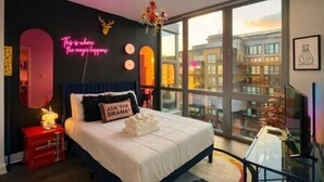 A modern bedroom with a large bed, a city view, and a neon sign on the wall.