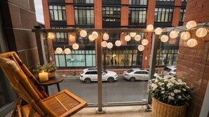 Uma varanda com móveis de vime, um cesto de vime com flores e vista para uma rua com carros estacionados.