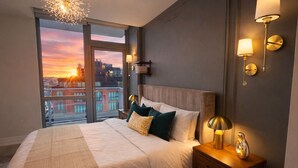 Une chambre d’hôtel moderne avec un grand lit, une table de chevet, une lampe et une vue sur la ville au coucher du soleil.
