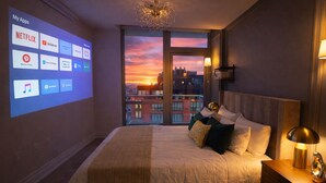 Une chambre d’hôtel avec un grand lit, une table de chevet, une lampe et une vue sur la ville au coucher du soleil.