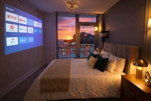 A hotel room with a large bed, a nightstand, a lamp, and a view of the city at sunset.