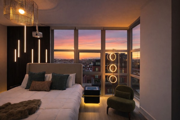 Et moderne soveværelse med en stor seng, et natbord, en stol og udsigt over byen i solnedgang.