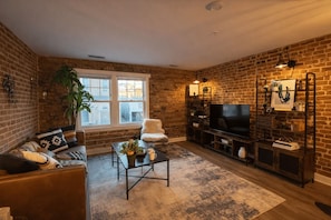 Uma sala de estar com parede de tijolos, uma cadeira branca, um sofá, uma mesa de centro, um aparador de TV e uma estante.
