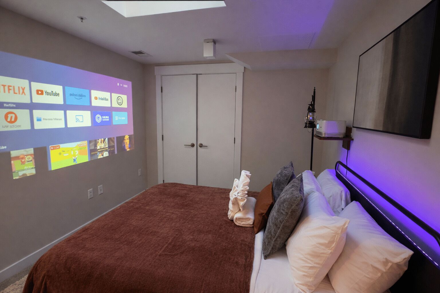 Ein Schlafzimmer mit einem Bett, Kissen, einem an der Wand montierten Fernseher, der verschiedene Streaming-Dienste anzeigt, und einer Deckenleuchte.