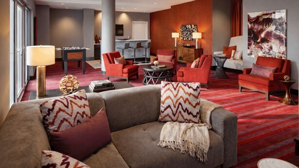 Een moderne hotellobby met een grijze bank, rode stoelen, een bar en een groot abstract schilderij.