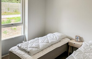 Un lit simple avec une couette et des oreillers blancs, une petite table de chevet sur laquelle est posée une photo encadrée, et une fenêtre donnant sur un paysage verdoyant.