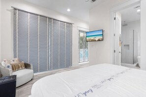 A modern bedroom with a large bed, a TV mounted on the wall, and a sliding glass door leading to a bathroom. - White Sands: Private Beach Access, HEATED Pool, Ocean Views, FREE Golf Cart (DESTIN)