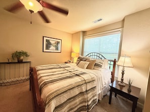 A bedroom with a bed, a ceiling fan, a window with blinds, a bedside table with a lamp, and a framed picture on the wall.