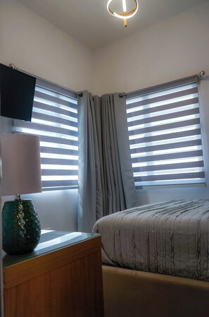 Un dormitorio con cama, televisor en la pared y ventana con cortinas.