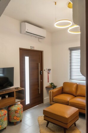 Una sala de estar moderna con un televisor de pantalla plana, un sofá de cuero marrón, un otoman a juego, un otoman con estampado floral, un mueble bajo de televisión de madera y un aire acondicionado instalado en la pared.