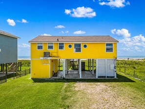 A two-story yellow house with a covered porch, a small attached garage, and a fenced yard. - Galveston Beachfront Home with Gulf Views, Beach Access and Waterfront Deck (Galveston)