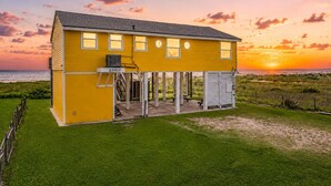A yellow building with a staircase leading to a covered area, a number sign, and a view of the ocean at sunset. - Galveston Beachfront Home with Gulf Views, Beach Access and Waterfront Deck (Galveston)