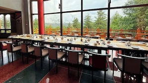 A dining area with a long table set for a meal, large windows with a view of greenery, and a red pillar. - Haven Resort Paro (Paro)