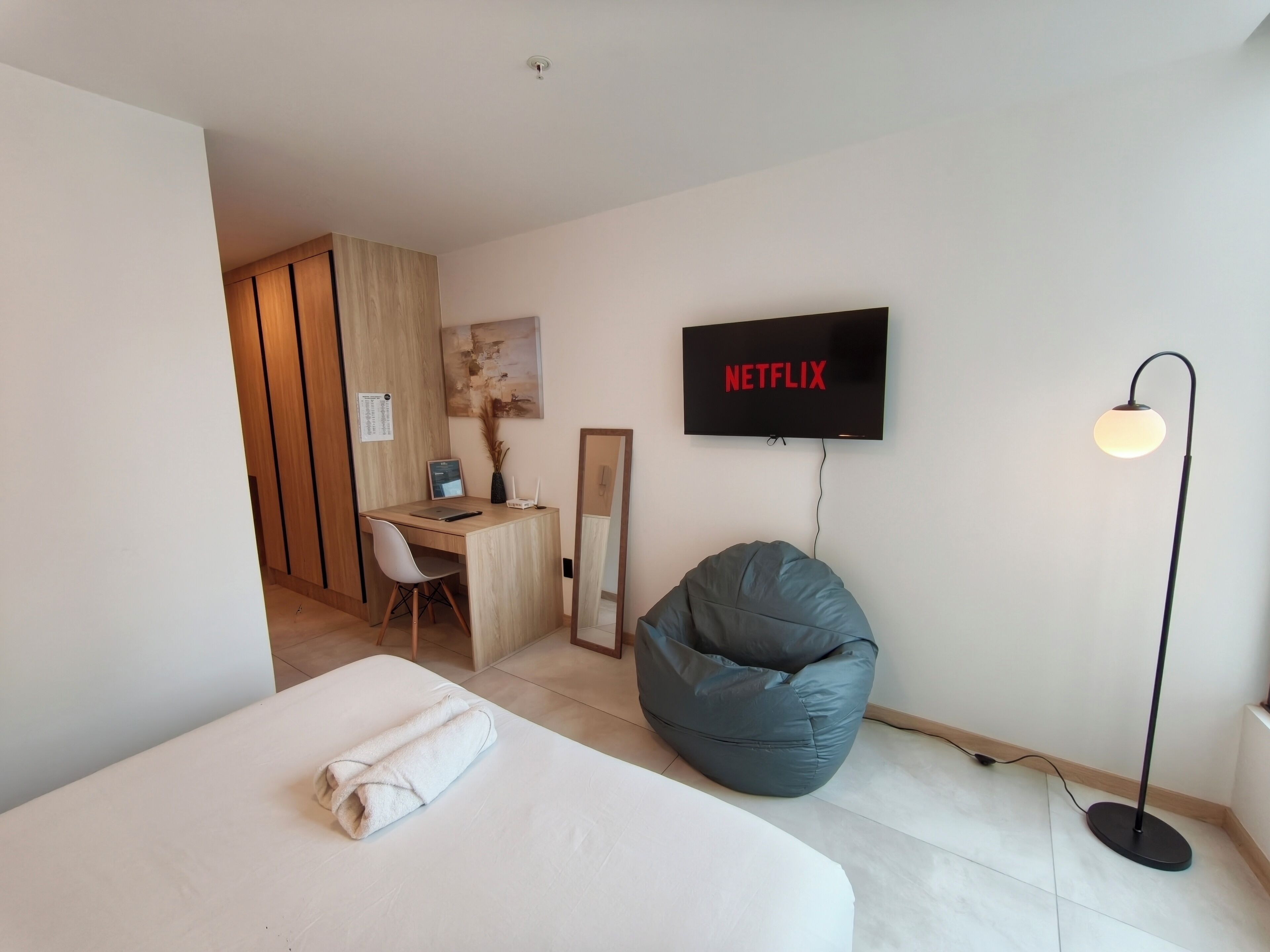 A modern hotel room with a bed, a desk, a chair, a bean bag, a TV, and a floor lamp.