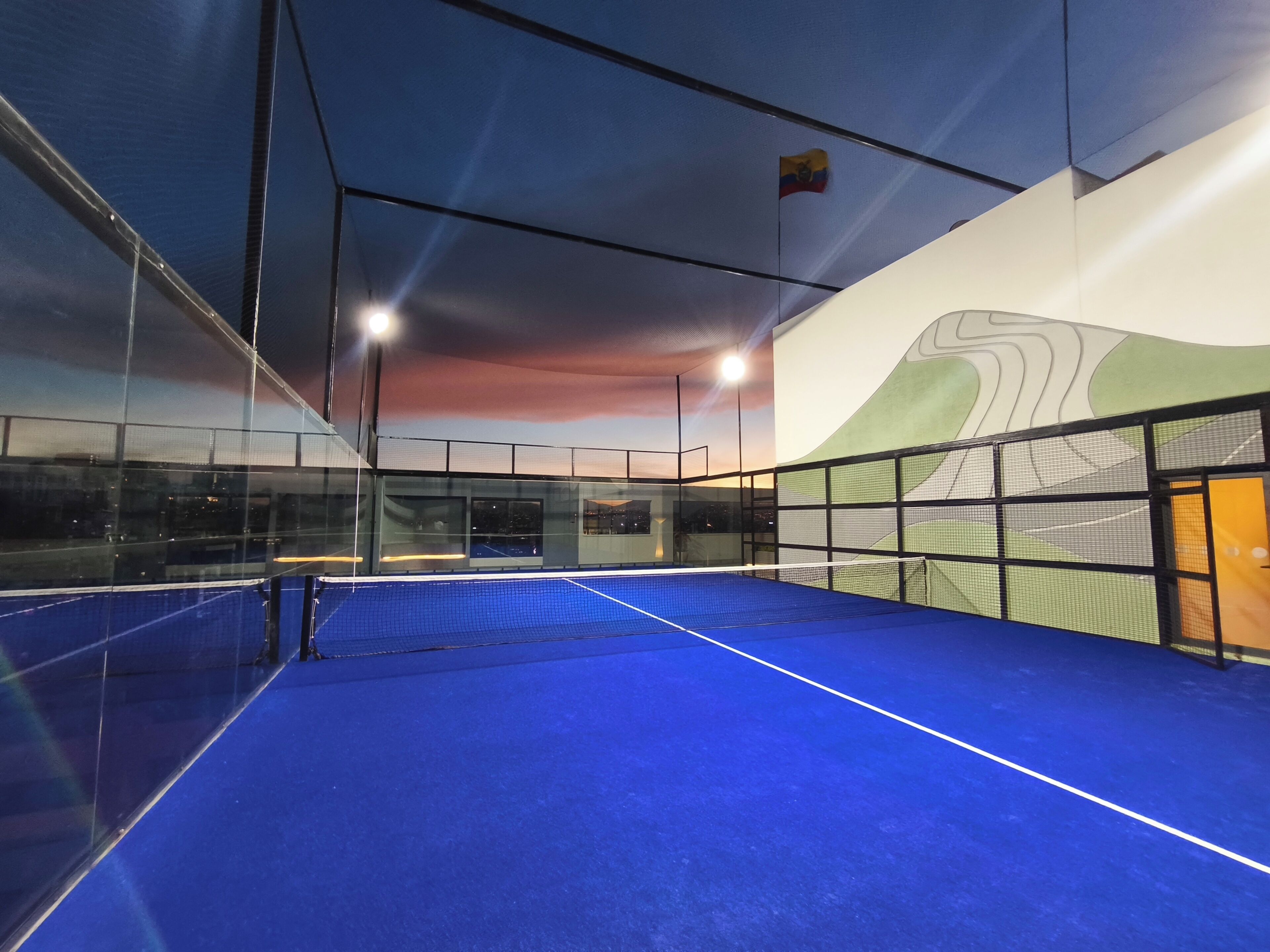 An indoor tennis court with a blue surface, glass walls, and a mural.